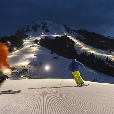 A nighttime skiing scene with two riders on the slope. In the background, the mountain shines with illuminated pistes.