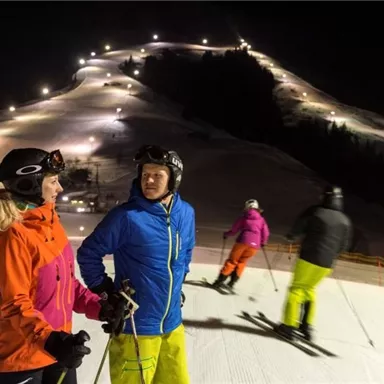 Two skiers are talking on a lit slope. In the background, more skiers and the snow-covered mountains can be seen.