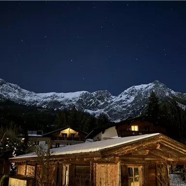 A picturesque mountain landscape at night with snow-covered peaks and a clear starry sky. In the foreground, a rustic wooden house can be seen.