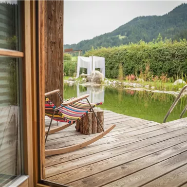 Ein gemütlicher Balkon mit einer Hängematte und Blick auf einen ruhigen Teich. Im Hintergrund sind sanfte Hügel und grüne Pflanzen zu sehen.