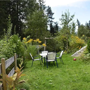 Ein gemütlicher Garten mit einem Tisch und Stühlen, umgeben von üppigem Grün und bunten Pflanzen. Im Hintergrund stehen hohe Bäume und ein klarer Himmel.