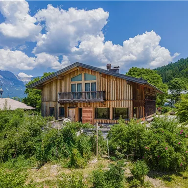 A beautiful wooden house surrounded by green plants and mountains. The sky is blue with white clouds.