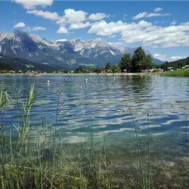 Ein klarer See umgeben von üppigem Grün und Bergen im Hintergrund. Der Himmel ist blau mit wenigen Wolken und es sind Menschen am Ufer sichtbar.