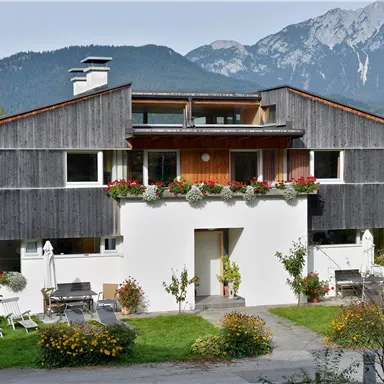 A modern house with a wooden facade and large windows. In the background, mountains and plants can be seen in a well-maintained garden.