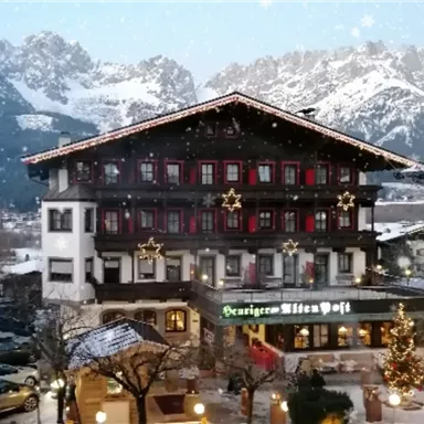 Ein traditionelles Hotel mit festlicher Weihnachtsdekoration in einer winterlichen Landschaft. Im Hintergrund sind majestätische Berge und ein klarer Himmel zu sehen.