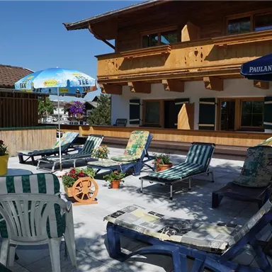 Eine gemütliche Terrasse mit Liegen und einem sonnigen Himmel. Im Hintergrund ist ein Holzgebäude mit Balkonen zu sehen.