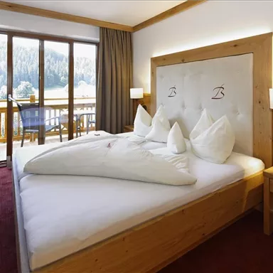 A cozy hotel room with a large bed and decorative pillows. The view through the windows showcases beautiful mountains and plenty of nature.