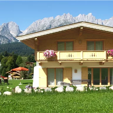 Ein charmantes alpines Haus mit einem Balkon und Blumenkästen. Im Hintergrund erheben sich majestätische Berge unter einem klaren blauen Himmel.