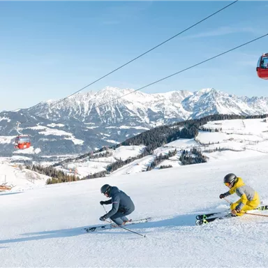 Zwei Skifahrer fahren die Piste hinunter, umgeben von schneebedeckten Bergen. Im Hintergrund sind Seilbahnen zu sehen.