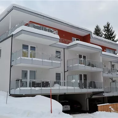 A modern apartment building in winter, surrounded by snow. The balconies are clearly visible and there are cars in the garage below.