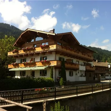 Ein malerisches Berghaus mit Holzbalcon und blühenden Pflanzen. Im Hintergrund sind grüne Berge und blauer Himmel zu sehen.