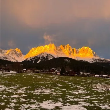 Eine beeindruckende Berglandschaft mit goldenem Licht auf den Gipfeln. Im Vordergrund sieht man eine grüne Wiese mit Schnee.