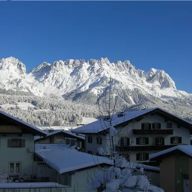 Eine schneebedeckte Berglandschaft mit hohen Gipfeln und klarem blauem Himmel. Im Vordergrund sind typische alpine Häuser zu sehen.