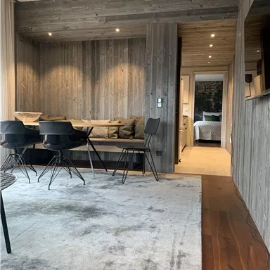 A modern living space with wooden paneling and a cozy seating area. In the background, a minimally designed sleeping area can be seen.