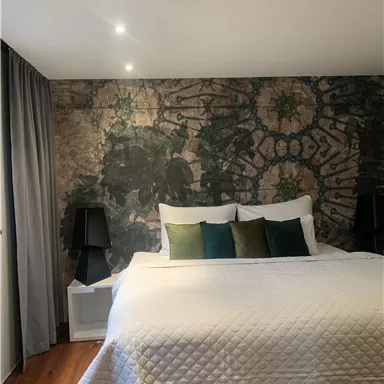 A stylish bedroom with a large bed and decorative pillows. The wall is adorned with a striking pattern, and there are two nightstands with lamps.