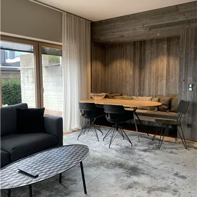 A modern living room with a black sofa and a round table. In the background, there is a wooden dining area with chairs.