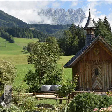 Eine charmante Holzkirche steht in einer grünen Landschaft mit Bergen im Hintergrund. Umgeben von Bäumen und Blumen blüht die Natur in voller Pracht.