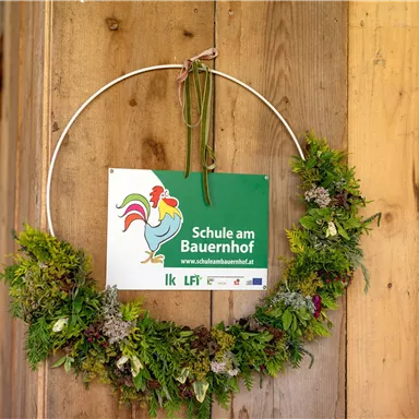 Ein grüner Kranz aus Pflanzen hängt an einer Holzwand. Darin befindet sich ein Schild mit der Aufschrift "Schule am Bauernhof".