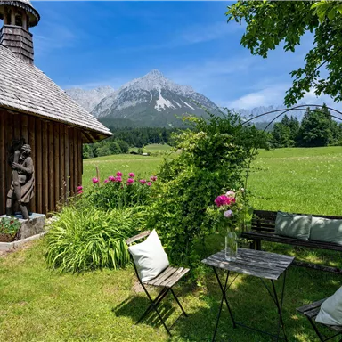 Eine gemütliche Ecke mit einem Tisch und Stühlen in der Nähe eines Holzhauses. Im Hintergrund sind grüne Wiesen und ein majestätischer Berg zu sehen.