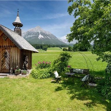 Eine kleine Holzkapelle steht in einer grünen Wiese, umgeben von Büschen und Blumen. Im Hintergrund sind majestätische Berge und ein klarer, blauer Himmel zu sehen.