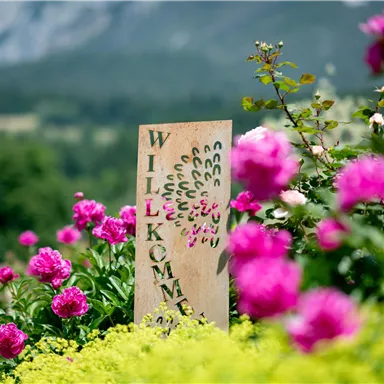 Ein Willkommenstein steht zwischen bunten Blumen. Im Hintergrund sind sanfte Hügel und grüne Landschaften zu sehen.