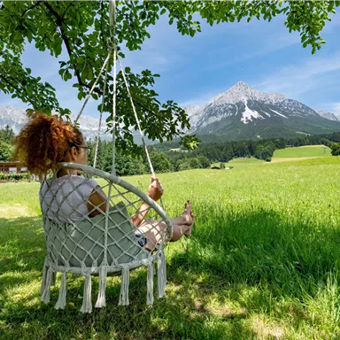 Eine Frau schwingt entspannt auf einer hängenden Schaukel in einer grünen Wiese. Im Hintergrund sind schneebedeckte Berge und ein blauer Himmel zu sehen.