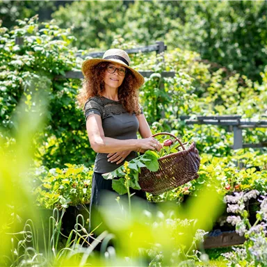 Eine Frau mit lockigem Haar und einem Hut steht in einem Garten und hält einen Korb mit frischen Pflanzen. Der Garten ist üppig und grün, umgeben von vielen Pflanzen.