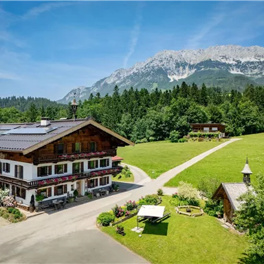 Ein malerisches Landhaus umgeben von grünen Wiesen und Bergen. Der Himmel ist klar und sonnig.