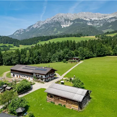 Eine malerische Landschaft mit sanften grünen Wiesen und bewaldeten Bergen im Hintergrund. Im Vordergrund sind zwei Gebäude zu sehen, die harmonisch in die Natur integriert sind.