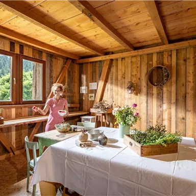 Ein gemütlicher Raum mit Holzwänden und einem gedeckten Tisch. Eine Frau in einem rosa Kleid steht beim Tisch und hält eine Schüssel.