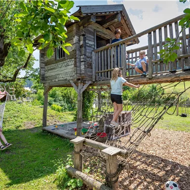 Ein Spielplatz mit einem Baumhaus und einer Kletterwand. Kinder spielen im Freien und genießen die Sonne.