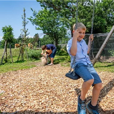 Ein Junge schwingt fröhlich auf einer Schaukel im Garten. Im Hintergrund spielen zwei Personen mit einem Haustier.