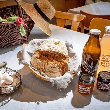 Ein rustikaler Tisch mit frischem Brot, zwei Flaschen Getränken und Marmelade. Im Hintergrund sind eine Kaffeetasse und ein Korb mit frischen Eiern und Gemüse zu sehen.