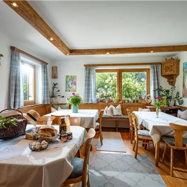 Ein gemütlicher Essraum mit hellen Tischen und Holzstühlen. Große Fenster bieten einen Blick ins Grüne und gestalten den Raum hell und einladend.