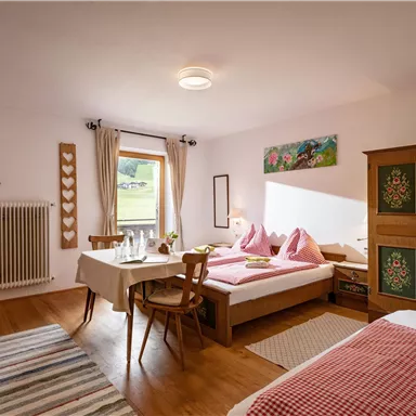 Ein gemütliches Zimmer mit zwei Betten, einem Tisch und Stühlen. Der Raum ist hell und freundlich, mit einem Fenster, das den Blick ins Freie ermöglicht.