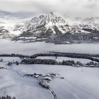 Eine schneebedeckte Landschaft mit hohen Bergen im Hintergrund. Der Himmel ist bewölkt und es gibt Nebel über den Feldern.