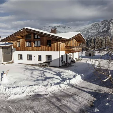 Ein gemütliches Ferienhaus in den Bergen, umgeben von frisch gefallenem Schnee. Im Hintergrund sind majestätische Berge und ein klarer Himmel zu sehen.
