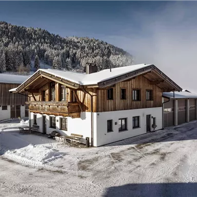 Ein traditionelles Holzhaus in einer schneebedeckten Landschaft. Im Hintergrund sind bewaldete Berge zu sehen.
