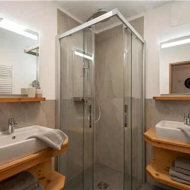 Ein modernes Badezimmer mit einer Glasdusche und zwei Waschbecken. Holzregale und Pflanzen sorgen für eine gemütliche Atmosphäre.