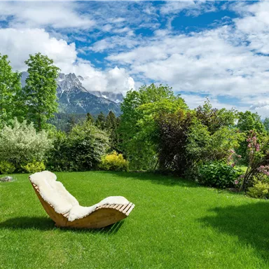 Ein schöner Garten mit sattgrünem Gras und bunten Pflanzen. Im Hintergrund sind beeindruckende Berge und ein blauer Himmel mit Wolken zu sehen.