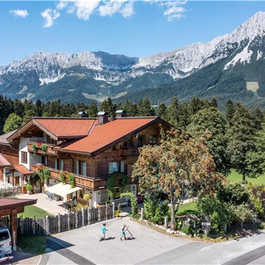 Ein traditionelles Alpenhaus umgeben von Bäumen und Wiesen. Im Hintergrund erheben sich beeindruckende Berge unter einem blauen Himmel.