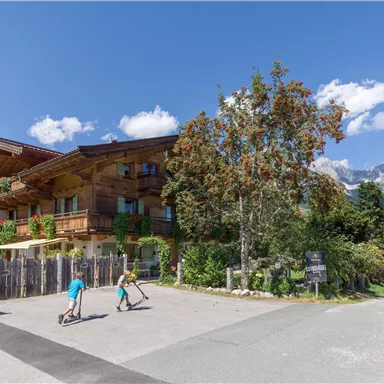 Ein charmantes Holzgebäude befindet sich in einer malerischen Landschaft. Zwei Kinder fahren mit ihren Rollern entlang einer Straße vor dem Gebäude.
