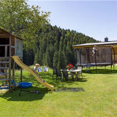 Ein Spielbereich mit einem Klettergerüst, einer Rutsche und einem Trampolin. Im Hintergrund sind Bäume und ein schönes Haus zu sehen.