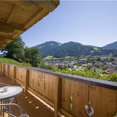 A balcony overlooking picturesque mountains and a small town. The sun is shining and the sky is clear blue.