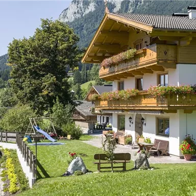 Ein gemütliches Haus mit einem schönen Balkon und blühenden Blumen. Der Garten ist grün und einladend, umgeben von Bergen und einer ländlichen Landschaft.