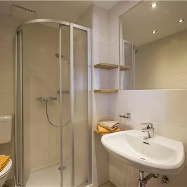 Ein modernes Badezimmer mit einer Duschkabine und einem Waschbecken. Es sind einige Handtücher und die Beleuchtung ist hell.