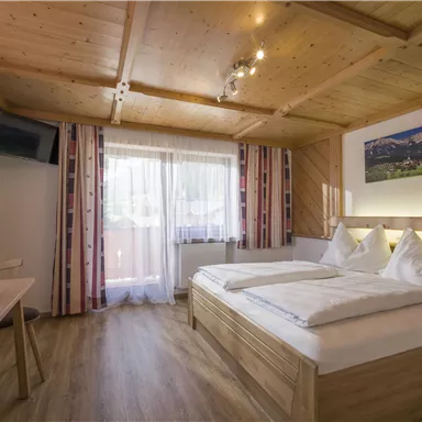 Ein gemütliches Zimmer mit Holzmöbeln und großen Fenstern. Auf dem Tisch steht ein Obstkorb, und es gibt einen Fernseher an der Wand.