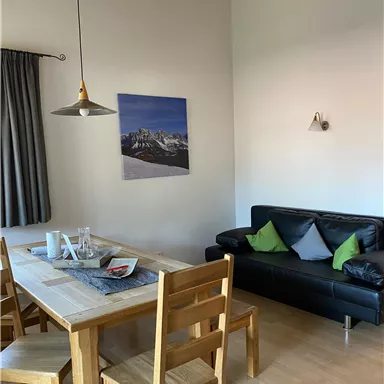 Ein gemütlicher Wohnraum mit einem Esstisch aus Holz und einer schwarzen Couch. An der Wand hängt ein Bild von Bergen, und die Fenster sind mit grauen Vorhängen bedeckt.