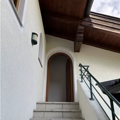 Eine Treppe führt zu einem Eingang unter einem Holzüberhang. Die Wände sind hell und die Tür ist architektonisch ansprechend.