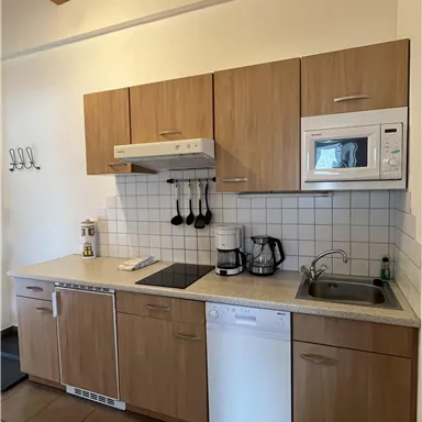 Eine moderne Küche mit Holzfronten und hellen Fliesen. Sie ist ausgestattet mit Herd, Mikrowelle, Kaffeemaschine und Geschirrspüler.